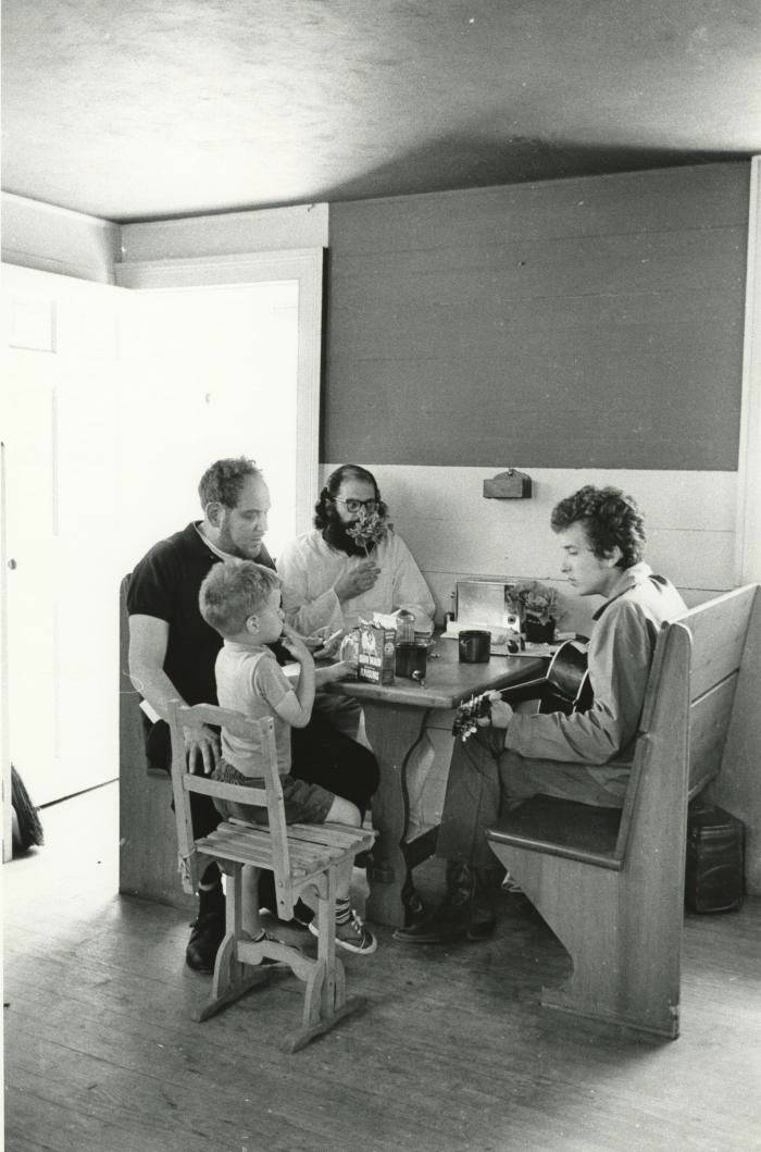 The photo is in black and white. At a table against the wall in a room sits Bob Dylan holding a guitar on his lap, and friends, Allen Ginsberg, Al Aronowitz and Son, w